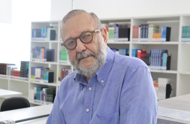 Retrato de homem de barba e óculos, camisa azul, em uma biblioteca com estantes de livros.