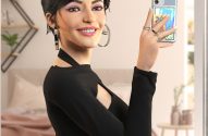 Mulher com cabelo preto e maquiagem rosa tira selfie no espelho do quarto.