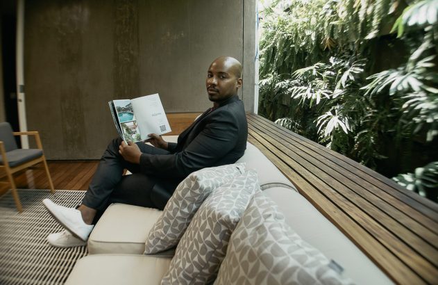 Um homem de terno sentado em um sofá lendo uma revista, olhando para a câmera. Ao fundo, uma parede verde com plantas e um deck de madeira.