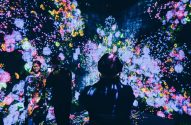 A imagem mostra pessoas em uma exposição de arte digital. O ambiente está escuro. A obra é exibida em telões e é composta por flores de diversos tipos e cores.
