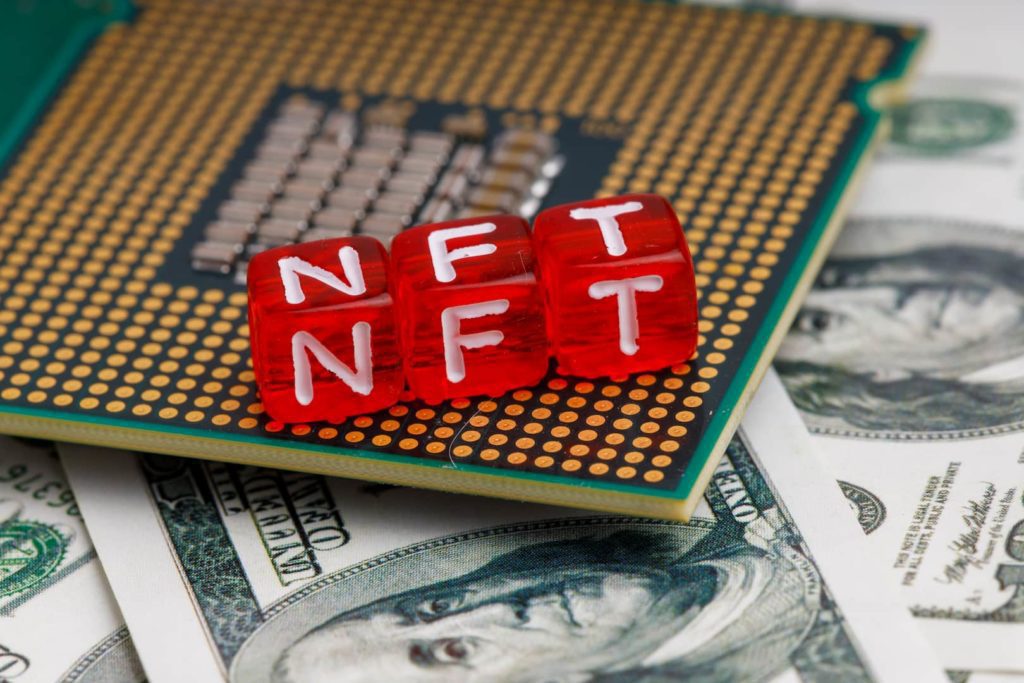 A imagem mostra três cubos vermelhos com letras, que formam a sigla NFT. Eles estão dispostos em uma placa de computador, que está em cima de várias notas de dólar.