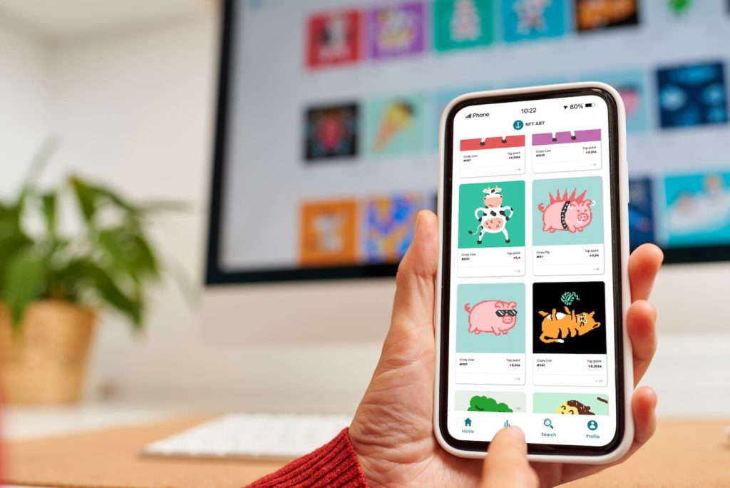 A imagem mostra um celular na mão de uma pessoa branca. Ela está acessando um marketplace com diversoas NFTs de animais. Ao fundo, que está desfocado, há um monitor que mostra o mesmo marketplace.