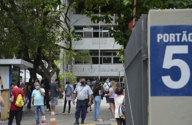 Pessoas com máscaras na entrada de um prédio universitário, com placa "PORTÃO 5" em primeiro plano.