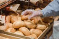 Mão com luva transparente pega pães frescos de um cesto de madeira em padaria/mercado.