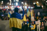Pessoa de costas com bandeira da Ucrânia em protesto noturno. Multidão com bandeiras ucranianas ao fundo.