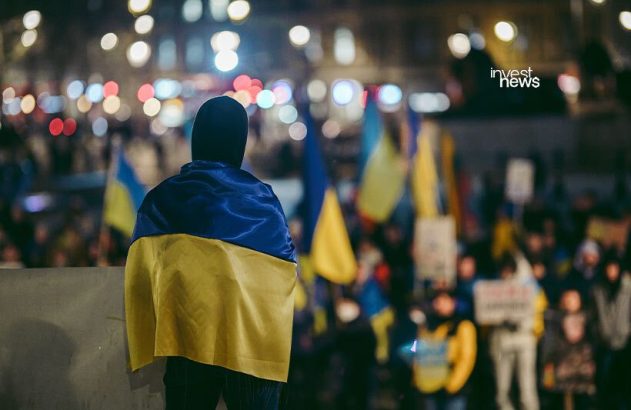 Pessoa de costas com bandeira da Ucrânia em protesto noturno. Multidão com bandeiras ucranianas ao fundo.
