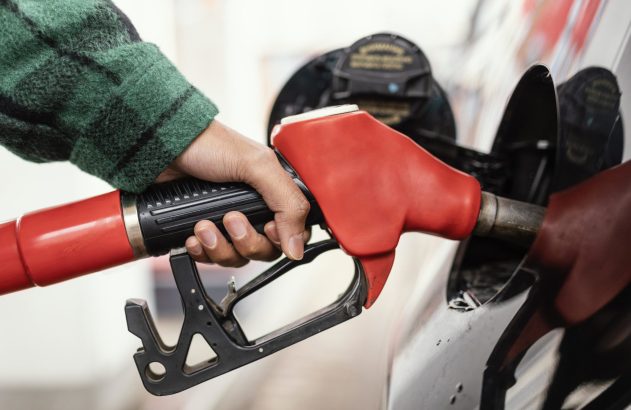 Mão segurando um bico de bomba de combustível vermelho, abastecendo um carro.