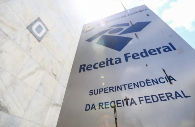 Sinal da Receita Federal com edifício de mármore e céu azul ao fundo.