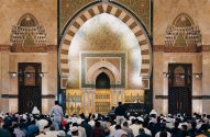 Fiéis em oração ou ouvindo sermão em mesquita. Interior ornado com arcos, mihrab e caligrafia islâmica.