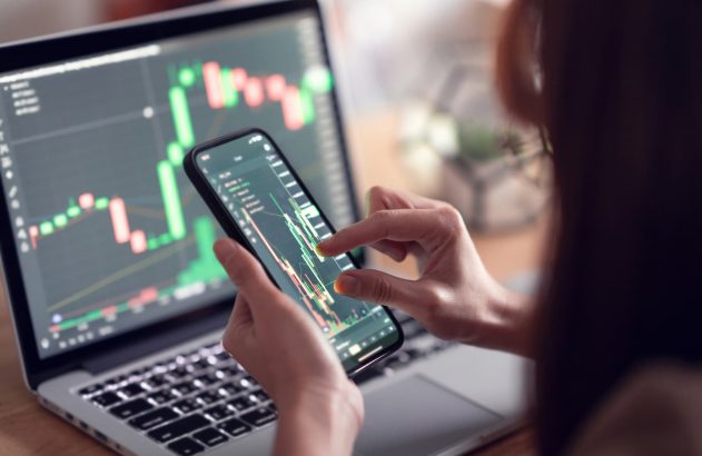 A imagem mostra uma mulher branca conferindo gráficos de cotação em um celular e em um notebook.