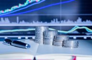 Pilhas de moedas, caneta e caderno aberto com gráficos financeiros em tela. Tema de investimento e análise de dados.
