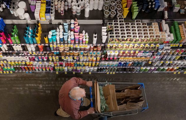 Vista superior de homem idoso empurrando carrinho de compras em loja. Prateleiras exibem diversos produtos.