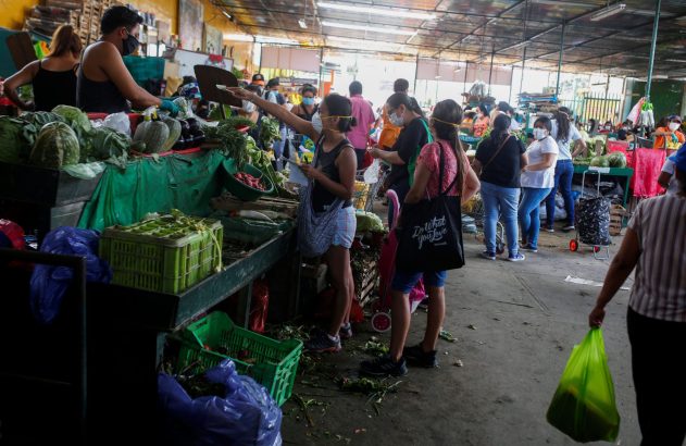 Mercado movimentado com pessoas comprando produtos frescos. Muitos clientes e vendedores usam máscaras faciais.