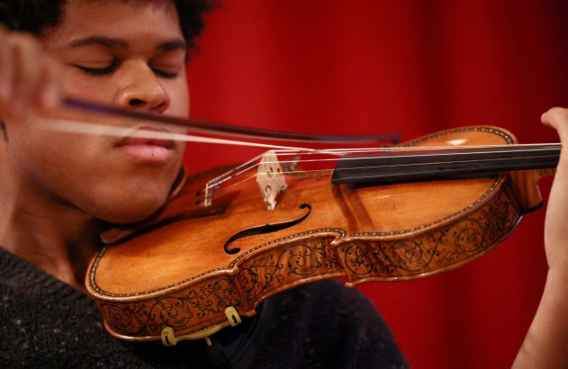 Jovem de olhos fechados tocando violino ricamente decorado. O arco está em movimento sobre as cordas. Fundo vermelho.
