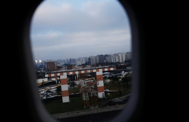Vista de janela de avião: cidade com prédios e estrutura quadriculada laranja e branca no aeroporto.