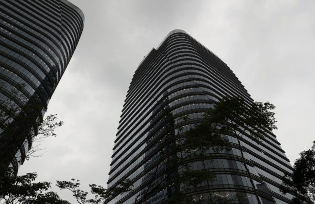 Dois edifícios altos de vidro curvo se elevam contra um céu cinzento, com árvores abaixo.