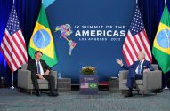 Líderes Biden e Bolsonaro sentados em reunião com bandeiras EUA/Brasil. Biden gesticula; Bolsonaro olha para baixo.