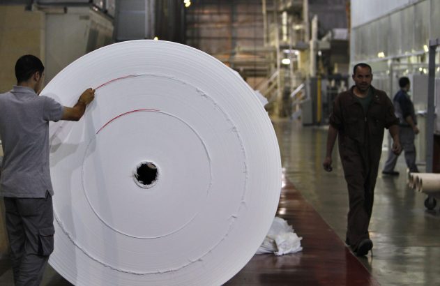 Trabalhador inspeciona enorme rolo de papel branco com marcas vermelhas, enquanto outro anda em chão de fábrica industrial.