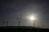 Silhuetas de torres de transmissão de energia contra o céu com um sol brilhante à direita, linhas de energia cruzam a imagem.