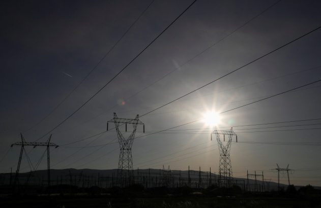 Silhuetas de torres de transmissão de energia contra o céu com um sol brilhante à direita, linhas de energia cruzam a imagem.