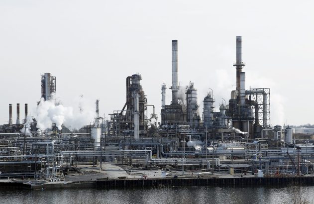 Refinaria industrial grande com chaminés e tubulações complexas, emitindo vapor. Um rio em primeiro plano.