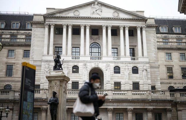 Banco da Inglaterra, fachada clássica com estátua de leão. Pessoa mascarada no primeiro plano, olhando celular.