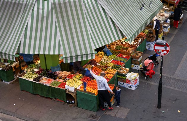 Mercado movimentado visto de cima, com toldo listrado, farta exibição de frutas e vegetais, e pessoas.