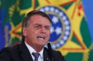 Jair Bolsonaro, de terno, com a boca aberta e um olho semicerrado, fala em microfone. Bandeira do Brasil desfocada ao fundo.