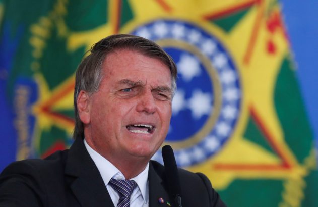 Jair Bolsonaro, de terno, com a boca aberta e um olho semicerrado, fala em microfone. Bandeira do Brasil desfocada ao fundo.