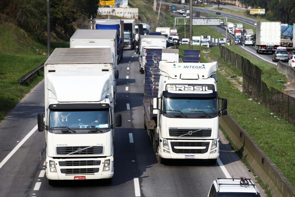 Longa fila de caminhões brancos em trânsito intenso numa rodovia, vistos de cima.