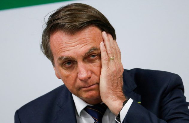 Jair Bolsonaro, de terno, com a mão esquerda na lateral da cabeça, olhando seriamente.