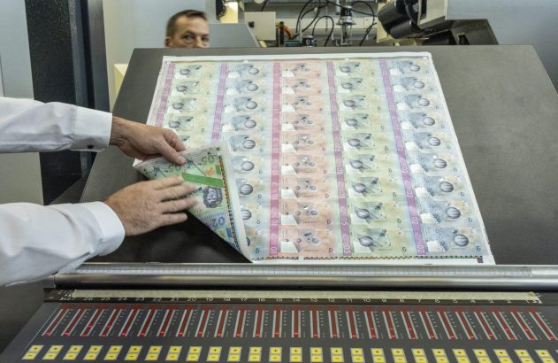 Mãos inspecionam folha de cédulas recém-impressas em uma impressora, com painel de controle e pessoa ao fundo.