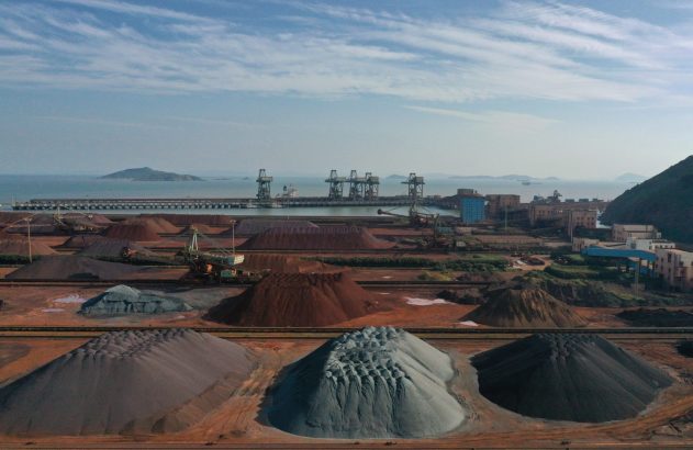 Vista aérea de porto industrial com extensas pilhas de minério, guindastes e navios em um terminal de carga a granel.