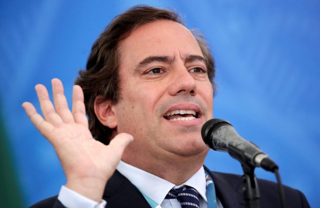 Homem de terno e gravata, com a boca aberta e a mão direita levantada, discursa em um microfone. Fundo azul.