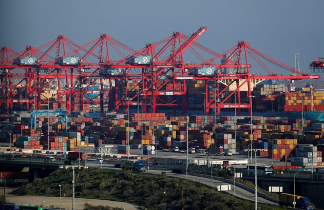 Guindastes vermelhos movimentam contêineres em um porto de carga, com pilhas e caminhões na rodovia.
