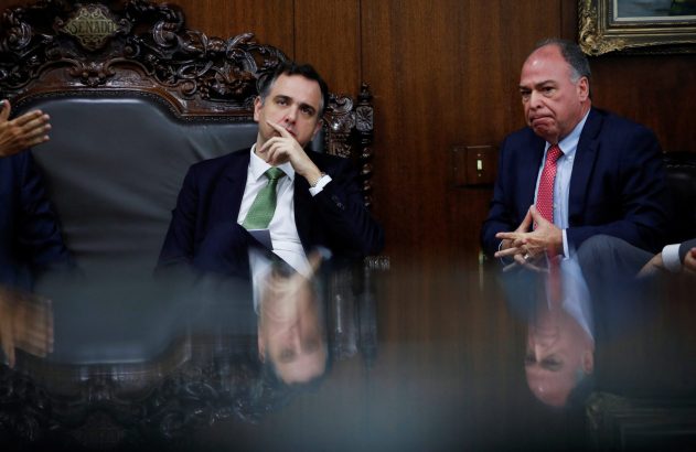 Dois homens de terno em reunião formal no Senado. Um pensativo, outro sério, com seus reflexos visíveis na mesa polida.