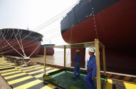 Dois grandes navios em estaleiro; trabalhadores no cais. Um navio tem bandeiras de inauguração.
