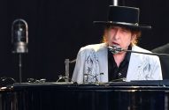 Bob Dylan de chapéu e jaqueta bordada canta e toca piano em palco.