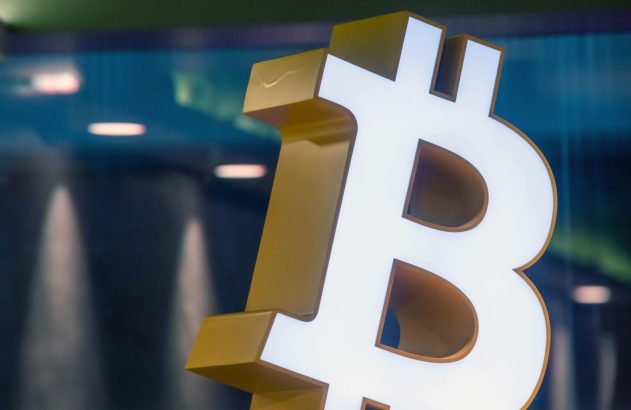 Logotipo 3D do Bitcoin branco e dourado, iluminado, contra um fundo azul escuro desfocado.