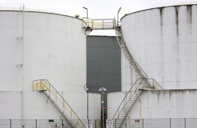 Dois tanques industriais brancos com escadas amarelas e passarela. Uma cerca em primeiro plano.