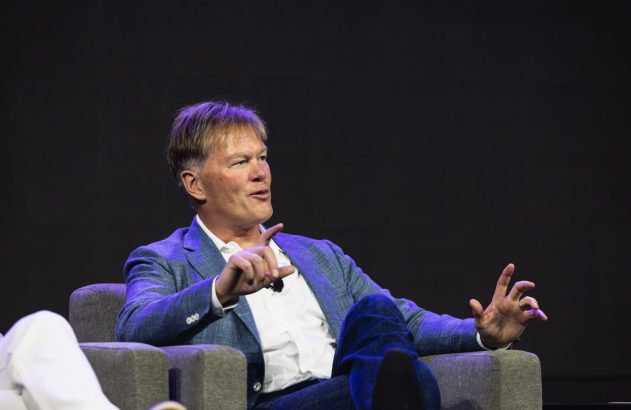 Homem de blazer azul e jeans, sentado em poltrona cinza, gesticula com as mãos em palco escuro.