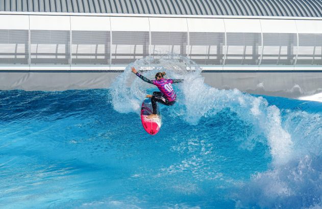 Surfista com roupa colorida e prancha rosa em piscina de ondas, criando grande spray de água na manobra.