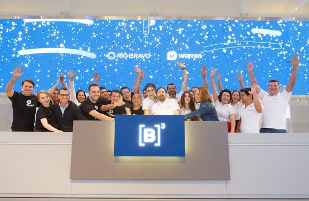 Grupo celebra com mãos para cima em evento da B3. Tela com logos Rio Bravo e Warren.