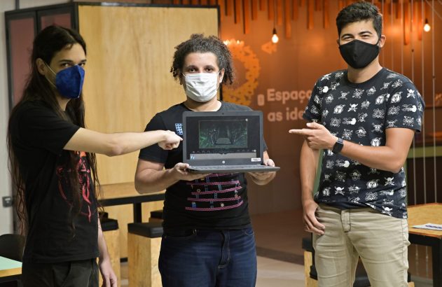 Três pessoas mascaradas apresentam um jogo em um laptop, enquanto duas apontam para a tela.
