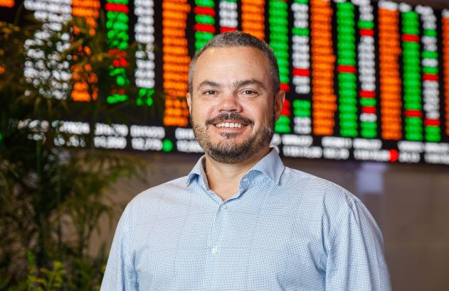 Homem sorrindo em frente a um painel de cotações financeiras.