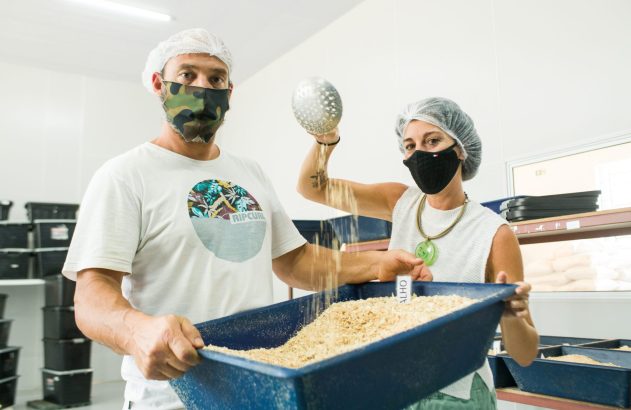 Duas pessoas com máscaras e toucas manuseiam bandeja de material granulado. Mulher adiciona pó com peneira. Rótulo "ALHO" visível.