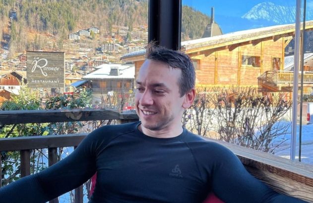 Homem sorridente sentado perto da janela, com vista para vila de montanha nevada e o restaurante 'La Rotonde'.