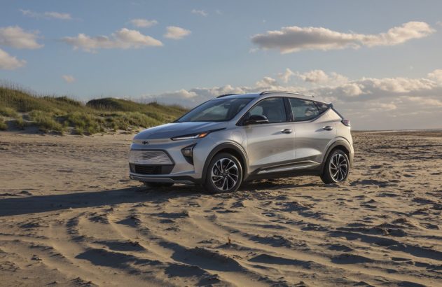 Chevrolet Bolt EUV prateado estacionado em praia arenosa com dunas e céu azul.