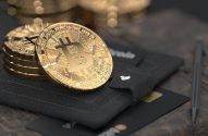 Moedas de Bitcoin douradas empilhadas, carteira escura com cartões e caneta sobre mesa marrom.