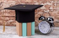 Capelo de formatura preto sobre livros e despertador em mesa de madeira.
